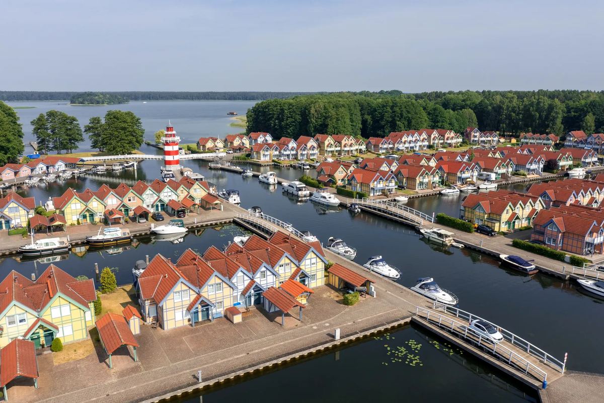 Luftbild: bunte Ferienhäuser an Hafenbecken mit Booten, dahinter ein See