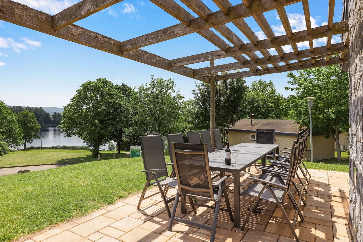 Terrasse mit Tisch unter einer Pergola, im Hintergrund ein See